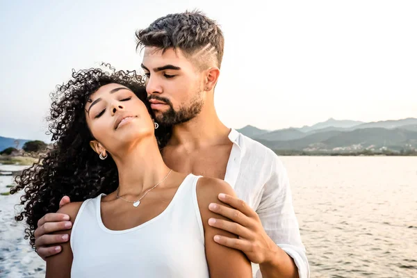 Heteroseksüel melez çift, gün batımında romantik bir sahnede klasik fotoğraf efekti filtresiyle yakışıklı beyaz erkek, İspanyol kız arkadaşını arkadan kucaklıyor ve gözleri kapalı, kıvırcık saçlara odaklan.