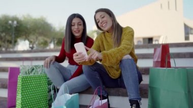 Kare basamaklarda oturmuş renkli çantalarla online alışveriş yapmaktan bahseden iki güzel kadın açık havada akıllı telefon kullanarak eğlenen bir çift kadın.