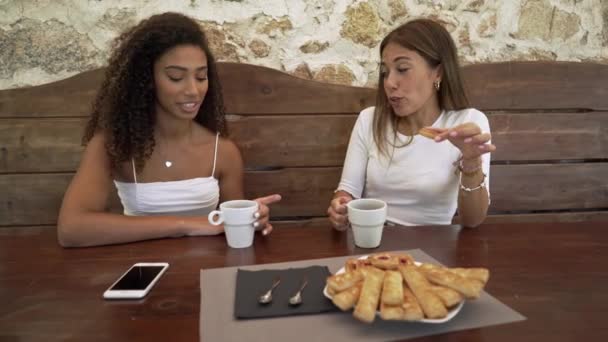 Weibliches Paar mit gemischter Rasse im Urlaub, das draußen am Holztisch einer Bar sitzt und Tee trinkt - Kaukasierin verbindet sich mit ihrer schwarzen hispanischen besten Freundin mit Gebäck und einer Tasse Kaffee