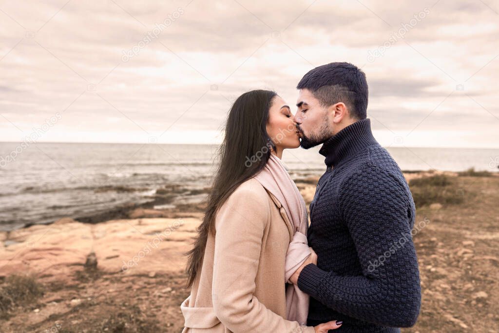 Mixed race couple romance scene on sea rocks and cloudy sky at sunset or dawn - Handsome bearded guy kisses his beautiful black Hispanic long hair woman holding her by the scarf with autumn colors