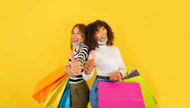 Renkli alışveriş torbaları tutan ve gülümseyen iki kendine güvenen melez genç kadın büyük sarı arka plan fotokopi alanındaki kameraya bakıp Coronavirus koruma maskesi takıyorlar.