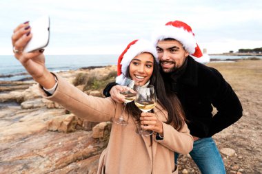 Genç, çok kültürlü bir çift, Noel Baba şapkası takıp, Noel tatilini ve kış tatilinde yeni yılı kutlayan beyaz şarap kadehleriyle deniz kenarında kendi portresini yapıyorlar.