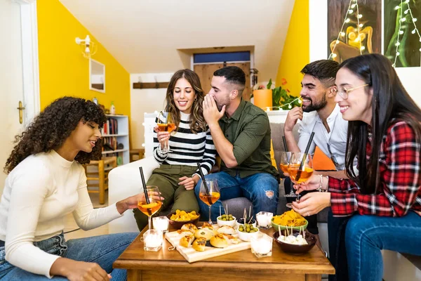 Group of mixed-race friends having fun talking to each other at home in the living room sitting on the sofa for a party drinking spritz long drink cocktail and various snacks on the coffee table