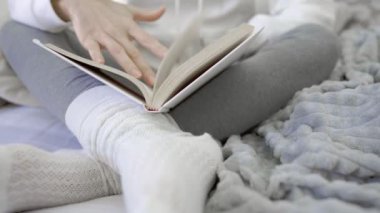 Kanepede oturan, bağdaş kurup beyaz yün çoraplara yaslanmış beyaz ve gri kış renklerinde metinleri takip eden bir kitap okuyan, tanınmamış genç bir kadının alt görünümü.