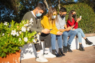Kentsel faaliyetlerdeki insanlar için yeni normal sosyal uzaklık: bin yıllık bir grup Coronavirus salgınına karşı koruyucu maskeli bir duvarda otururken dışarıda akıllı telefon kullanarak içeri girmek için bekliyor