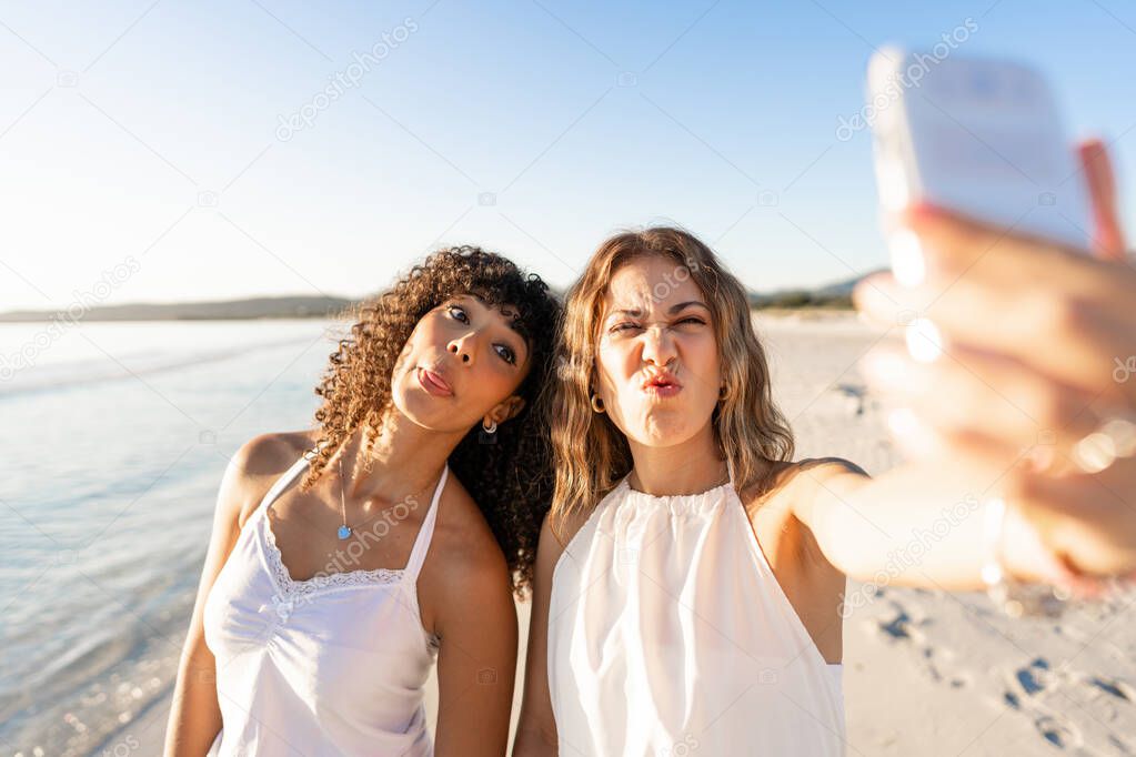 Beautiful mixed race female love couple making faces doing self portrait on beach - Two lesbian pretty women having fun using smartphone to sharing diversity - Selective focus on right girl face