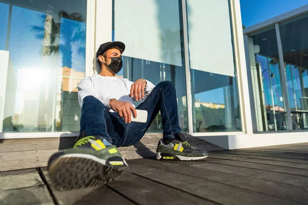 Young modern bored guy sitting on the ground in the city street leaning against a store window wearing Coronavirus protective mask, hat holding smartphone. Concept of new normal young people life