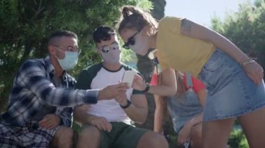 Coronavirus koruyucu maskesindeki şehir parkında akıllı telefon kullanan bir grup genç arkadaş. Sokağın ortasında cep telefonlu ve güneş gözlüklü iki çift internette eğleniyor.