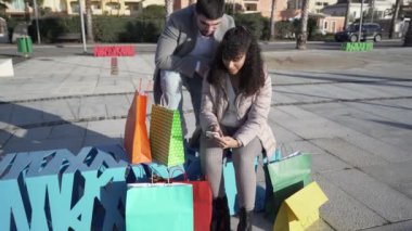 Genç, alışveriş düşkünü, çok kültürlü bir çift, renkli kağıt torbaların arasındaki online alışverişleri kontrol etmek için akıllı telefonlarıyla dışarıda eğleniyor. Uzak bağlantı nedeniyle yeni satın alma yolu