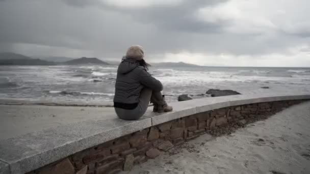 冬のドレスを着た無名の若い女性と 劇的な曇りの雨の空の下で嵐の海の波を見て後ろから撃たれた海岸沿いの壁に座っているウールの帽子 人は地平線を見る — ストック動画