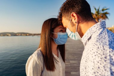 Coronavirus yüzünden koruyucu maske takarak birbirlerinin alnına yaslanarak birbirlerinin gözlerine bakarlar. Gün batımında ya da şafakta okyanus kıyısındaki tatil beldesinde salgın hastalıklı iki genç turist.