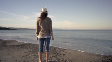 Arkadan vurulan tanınmayan genç turist kadın, Coronavirus 'a karşı koruyucu maskesini tutan ıssız bir sahilde denize bakıyor. Covid-19 turizme zarar verdi