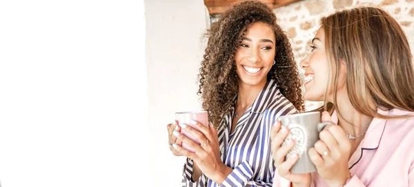 Multiracial women couple smiling looking each other holding a teacup in pajama just waken up. New normal gay families relationship and habits daily life scenes. Afro-American brunette curly woman