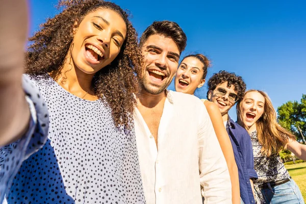 Five young multiracial friends in line posing for photo portrait smiling with open mouth. Black Hispanic brunette girl taking a selfie with smartphone outdoor. POV of happy millennial carefree people