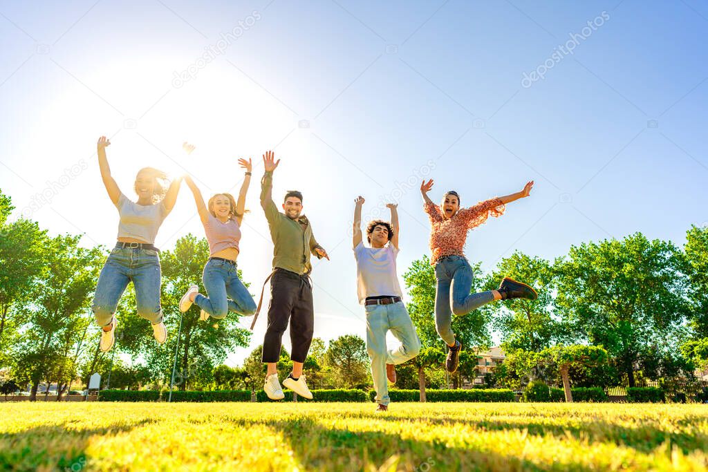 Backlit photography of multiracial group of college students jumping high with open up arms smiling looking at camera in a sunny day with blue sky at city park. Concept of better success in teamwork