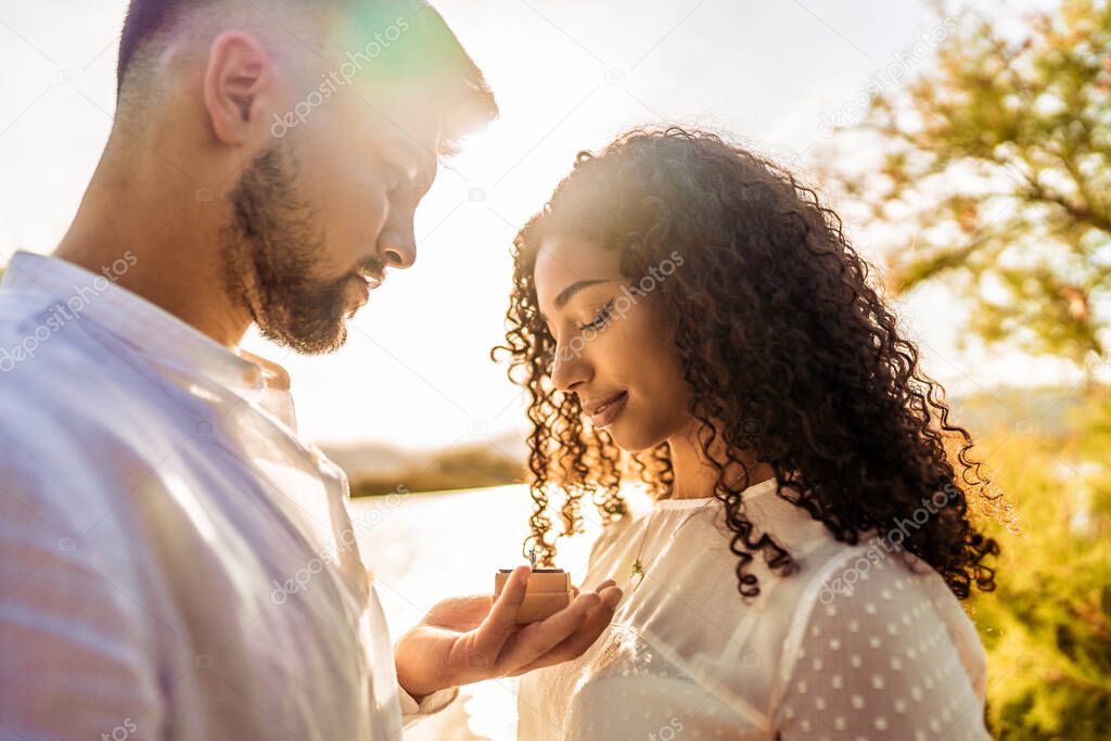 Backlit suggestive romance scene of young beautiful multiracial couple in love. Handsome guy making the marriage proposal to her black Hispanic girlfriend showing engagement ring close to her