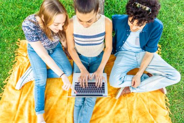 Şehir parkındaki bir ağacın altında dizüstü bilgisayarla oturan üç gen z öğrencisinden online dersler için arkadaşlarla bağlantı kurmaya kadar görüntüyü al. Gençler defter ve internetteki kablosuz bağlantılarla eğleniyor