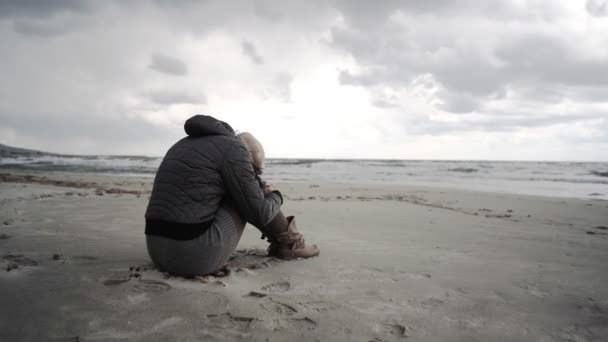 难以辨认的孤寂绝望的女人 在冬日的大海中 坐在沙滩上沉思 两腿夹着头 沉思着未来的命运 在灰蒙蒙的天空下 悲伤地回忆着过去 — 图库视频影像
