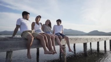 Two young multiracial smiling couple enjoying summer in nature sitting on wooden pier with dangling feet on flat water. Carefree friends spending time talking to each other at vacation resort at dusk