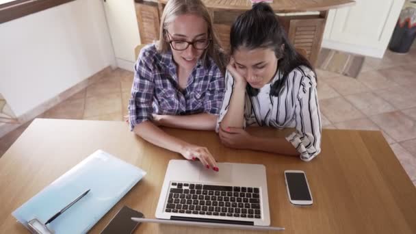 两个年轻的学生坐在客厅的桌子旁 在笔记本电脑和智能手机上工作 在网上学习 新的远距离正常的学校活动 使用无线移动技术的女孩 — 图库视频影像