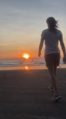 Günbatımını izlemek için Koh Lanta 'nın kumlu kıyılarında yürüyen genç bir kadının güzel dikey 4K 60fps görüntüleri. Onun silueti alevli gökyüzüne ve parıldayan okyanusa karşı huzur, gezgin şehvet ve doğayla bağlantıyı çağrıştırıyor..