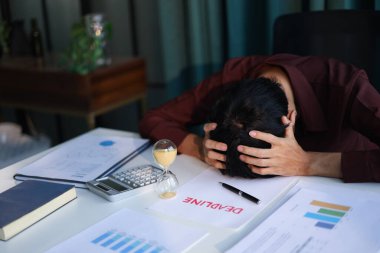 İş adamı başını tutuyor, uyuyor, ofis masasında son teslim tarihi belgeleriyle dinleniyor ve kum saati çalışıyor. Zaman yönetimi kavramı.