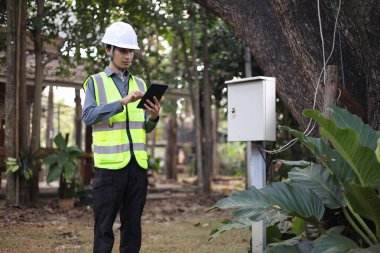 Mühendis ya da güvenlik uzmanı kişisel koruyucu giysiler giyiyor, dışarıdaki bir elektrik kutusunu denetlemek ya da yönetmek için tablet kullanıyor.