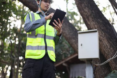 Mühendis ya da güvenlik uzmanı kişisel koruyucu giysiler giyiyor, dışarıdaki bir elektrik kutusunu denetlemek ya da yönetmek için tablet kullanıyor.