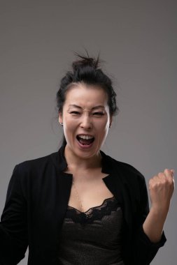 Enthusiastic motivated successful Asian business woman cheering and clenching her fist over a grey studio background with copyspace clipart