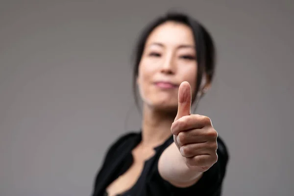 得意洋洋的亚洲女人举起大拇指以示成功和赞许 把注意力集中在拥有版权的灰色工作室背景上 — 图库照片