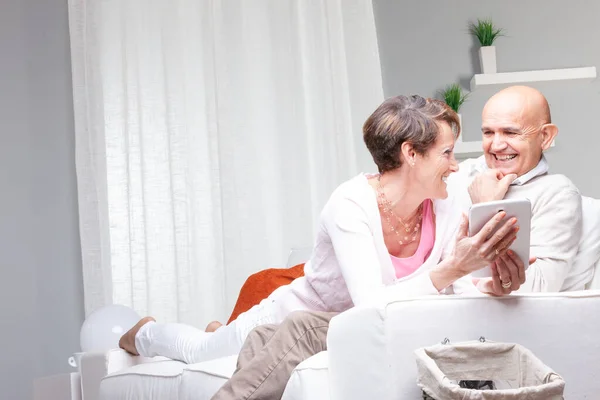 一对中年夫妇在平板电脑上对媒体笑得很开心 他们在家里沙发上放松的时候 面带微笑地看着对方 — 图库照片