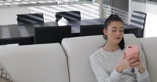 Молодая женщина с розовым телефоном на диване говорит — стоковое видео