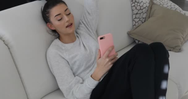 Mujer joven en el sofá con teléfono inteligente rosa - clip de material de archivo — Vídeos de Stock