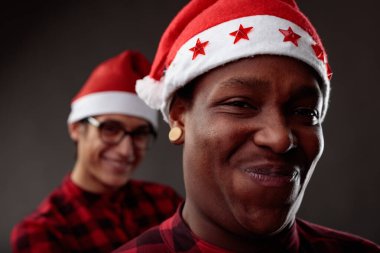 Charismatic male friends grinning at the camera in Santa Hats as they celebrate the Christmas festive holiday season with focus to a smiling Black man in the foreground clipart