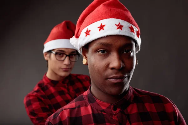 Playful young men friends wearing colorful red Santa Hats to celebrate the Christmas season smiling quietly at the camera as they mirror each others expressions in an over the shoulder view