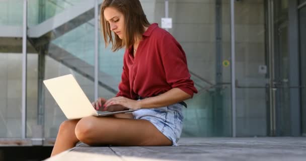 Young woman writing outside on a laptop in shorts — Stockvideo