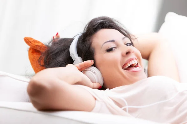 年轻而快乐的女人 在舒适的沙发上舒服地舒服地舒服地靠在近旁 一边用立体声耳机听着自己的音乐 一边高兴得很 — 图库照片