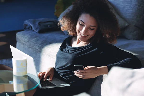 在Covid 19大流行期间 在家里的笔记本电脑上工作的年轻女性坐在地板上 开心地笑着在手机上留言 — 图库照片