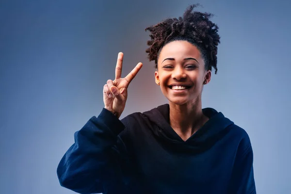 Studio portrait of an African American cheerful young woman showing victory sign while looking at camera against blue background for copy space