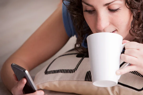 Menina aproveitando seu celular enquanto bebe de uma caneca — Fotografia de Stock