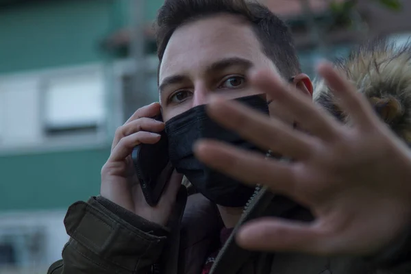 man with disposable surgical mask on his face for protection from viruses and infections making the stop sign with his hand while he talks on the phone