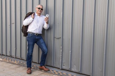 Yetişkin bir adam cep telefonu kullanarak duvara yaslanıyor.