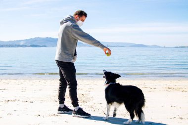 Tıbbi maskeli bir adam, köpeğiyle kumsalda oynuyor.