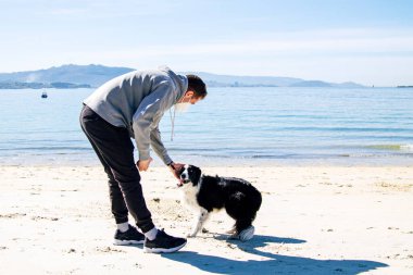 Tıbbi maskeli bir adam, köpeğiyle kumsalda oynuyor.