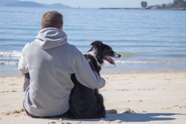 man sitting in front of the sea on the beach with his dog friend