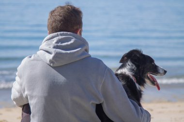 man sitting in front of the sea on the beach with his dog friend