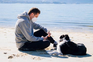 man with medical mask on the beach with his dog and using the phone