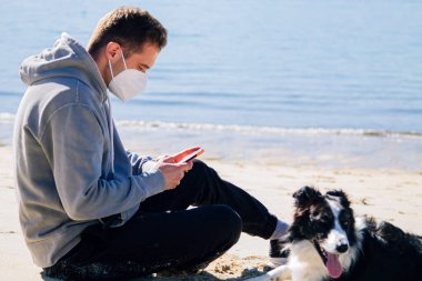 man with medical mask on the beach with his dog and using the phone