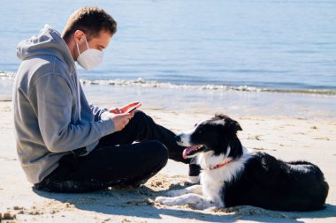 man with medical mask on the beach with his dog and using the phone