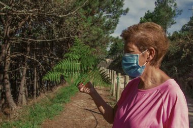 Tek kullanımlık tıbbi maskeli bir kadın ormanda eğrelti otu tutuyor.